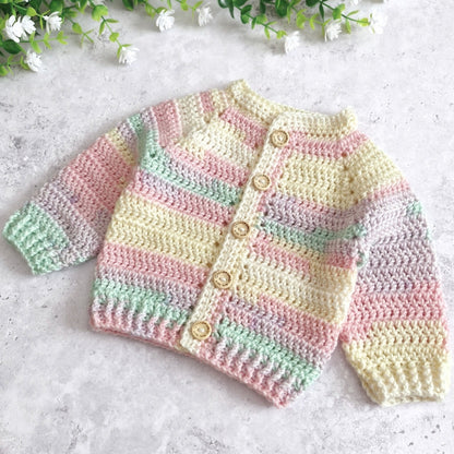 A pastel crocheted cardigan sweater for babies, featuring horizontal stripes of pink, mint green, and cream, with five wooden buttons down the front. It is displayed on a light gray textured surface with small white flowers at the top.