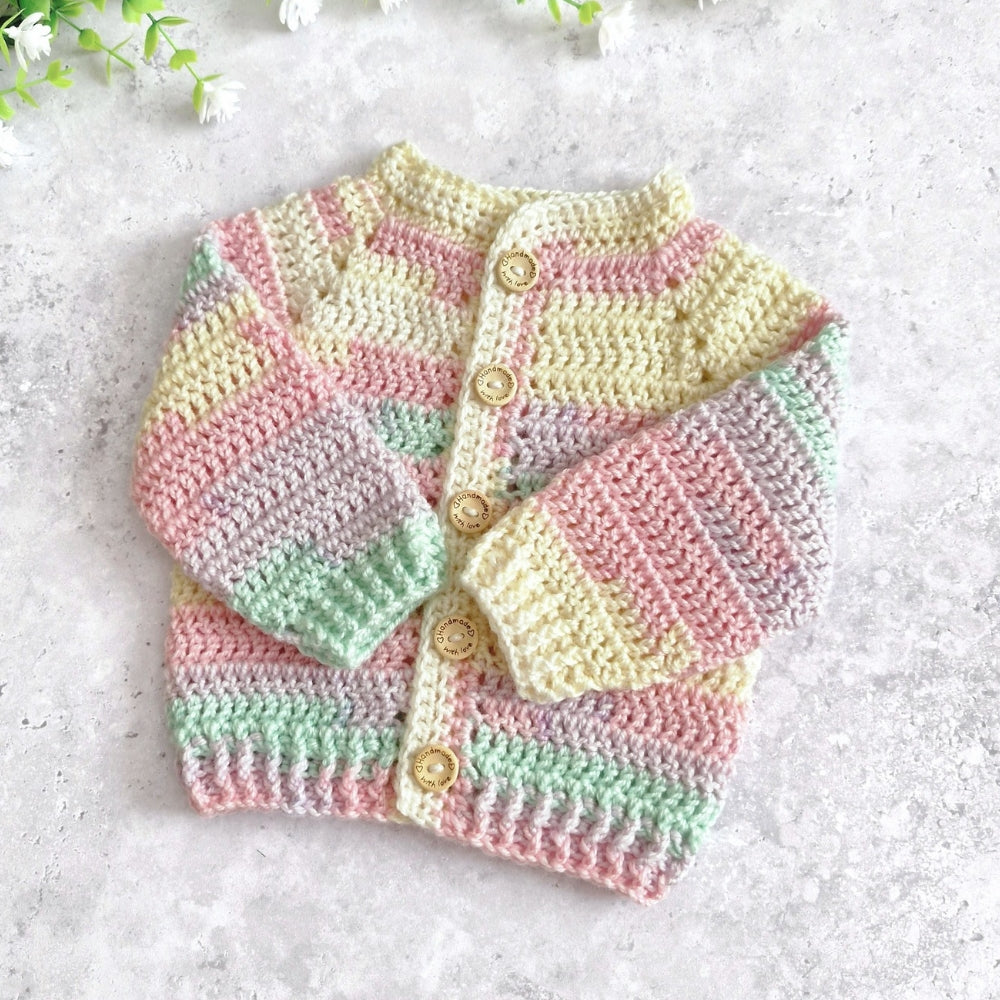 A pastel crocheted cardigan sweater with horizontal stripes in pink, yellow, green, and lavender, featuring wooden buttons, laid flat on a light gray textured surface.