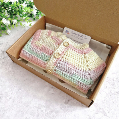 A pastel striped crocheted cardigan or sweater displayed inside an open brown cardboard box on a light surface, with a small card reading 'Twisted by the Sea' visible inside the box.
