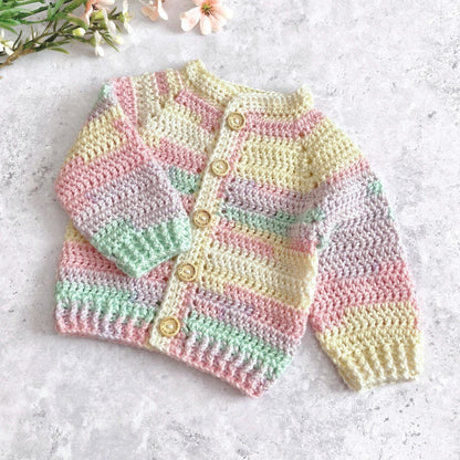 A handmade crocheted baby cardigan with pastel stripes in pink, yellow, green, and purple, featuring wooden buttons, laid flat on a light gray textured surface with small flowers in the corner.