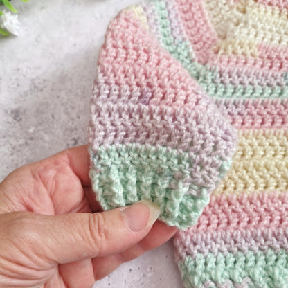 A close-up of a hand holding a soft crocheted sleeve, showing pastel stripes in pink, mint green, pale yellow, and off-white against a light stone-like background.