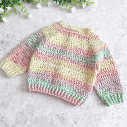 A back view of a pastel striped crocheted baby sweater with long sleeves, laid flat on a light gray textured surface with small white flowers at the top.