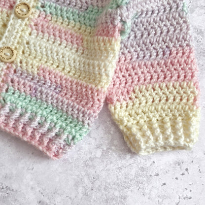 A close-up of a handmade crocheted cardigan with pastel stripes in pink, yellow, green, and lavender, featuring two wooden buttons on the left side, laid flat on a textured light gray surface.