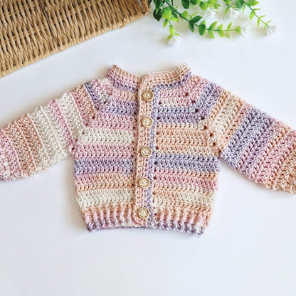 A handmade crocheted cardigan sweater for babies, featuring soft pastel stripes of pink, purple, beige, and white, with a button closure. It is laid flat on a white surface with a woven mat and green leaves in the background.