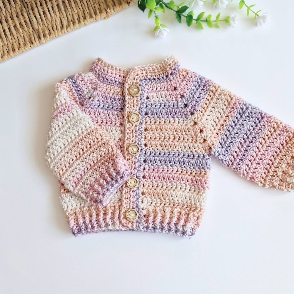 A handmade crocheted baby cardigan with pastel stripes in pink, purple, beige, and white, featuring a button-up front with wooden buttons, laid flat on a white surface.