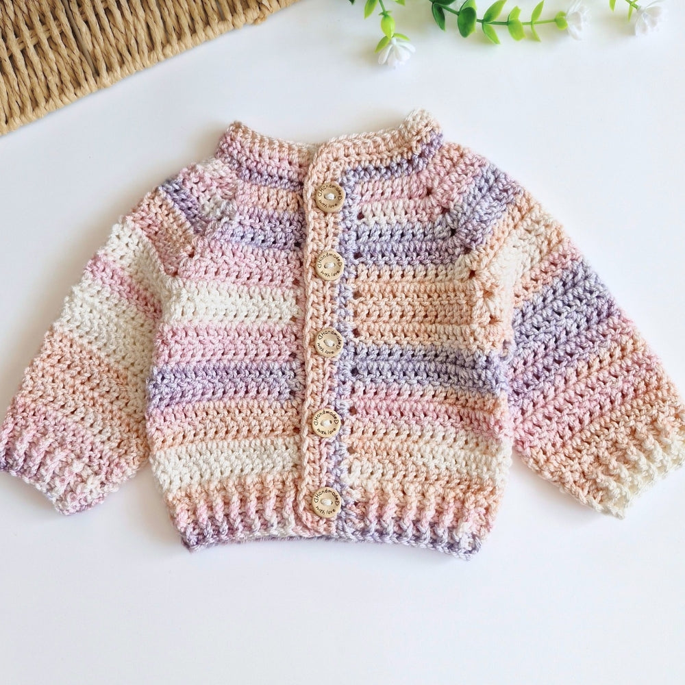 A handmade crocheted baby cardigan with horizontal stripes in pastel pink, cream, and lavender, featuring four wooden buttons down the front, laid flat on a white surface.
