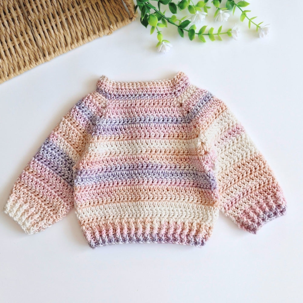 A handmade crocheted baby sweater with horizontal pastel stripes in pink, beige, and lavender, laid flat on a white surface with a woven mat and green leaves in the background.