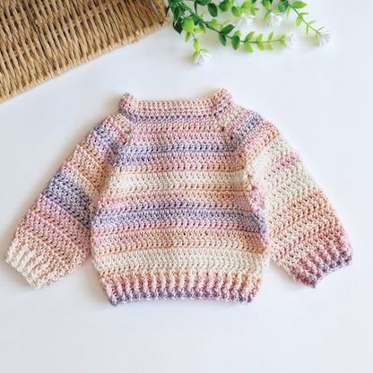 A handmade crocheted baby sweater with horizontal pastel stripes in pink, beige, and lavender, laid flat on a white surface with a woven mat and green leaves in the background.