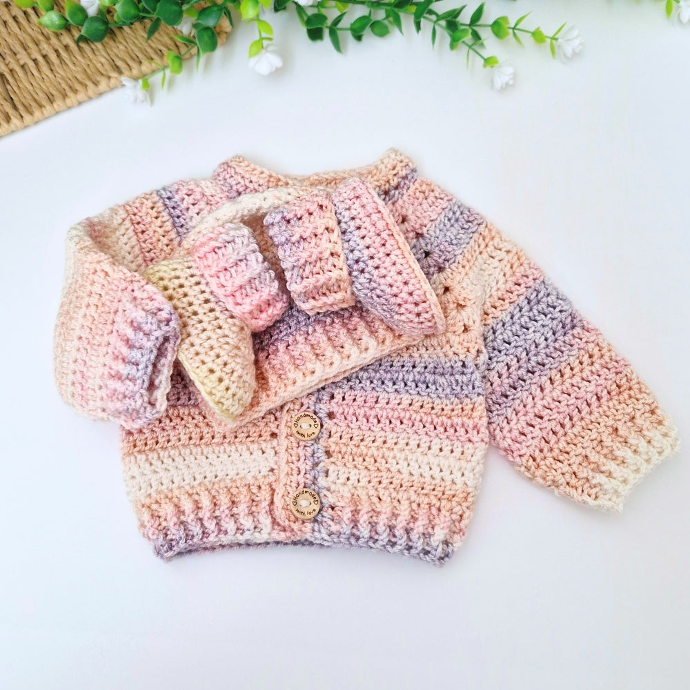 A handmade crocheted baby cardigan set with a matching hat and booties, featuring soft pastel stripes of pink, peach, beige, and lavender, styled on a white surface with decorative greenery.