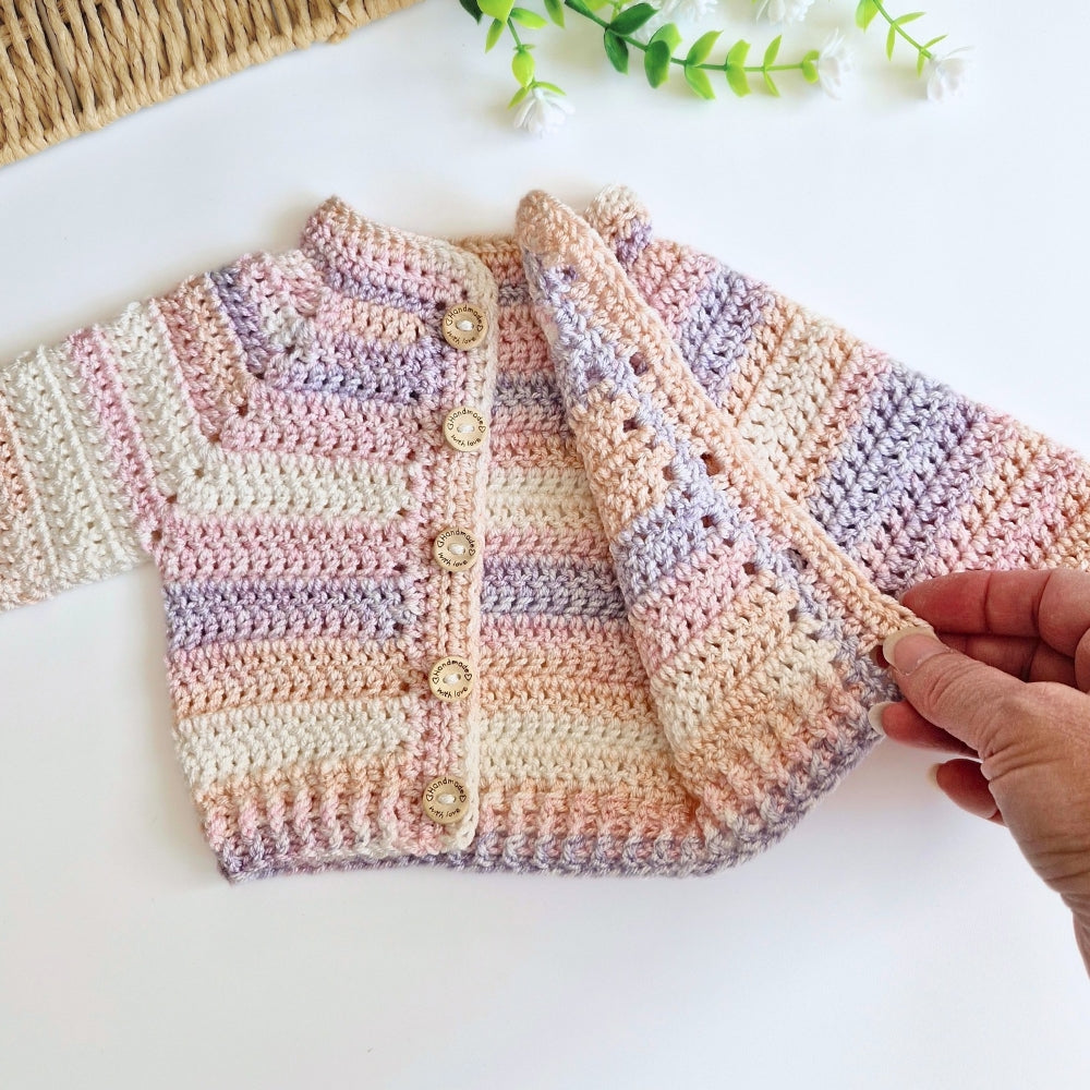 A hand-crocheted baby cardigan in pastel stripes of pink, beige, and lavender, featuring gold buttons, shown with a hand holding one side on a white surface.