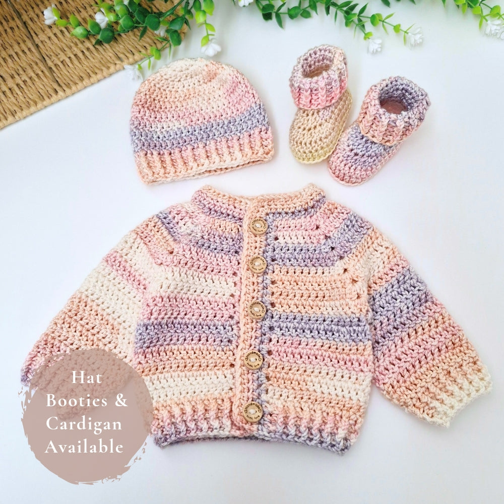 A handmade crochet baby outfit set laid flat on a white surface, including a cardigan, hat, and booties. The crochet pieces feature soft pastel stripes in pink, cream, and lavender-gray, with wooden buttons on the cardigan.