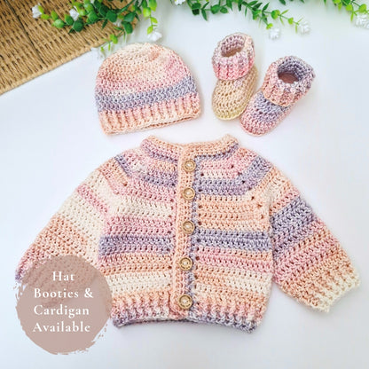 A handmade crochet baby outfit set laid flat on a white surface, including a cardigan, hat, and booties. The crochet pieces feature soft pastel stripes in pink, cream, and lavender-gray, with wooden buttons on the cardigan.