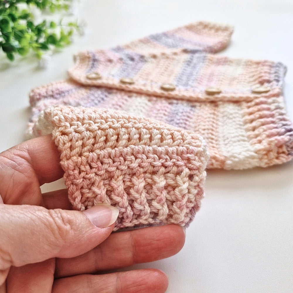 A close-up of a hand holding a small crocheted pouch or card holder made from soft pastel yarn, with a larger matching pouch laid out in the background on a light surface.
