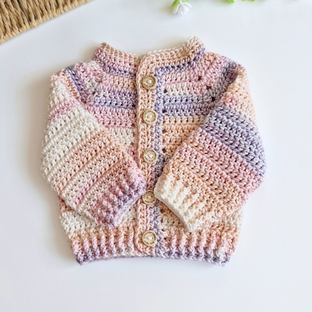 A handmade, long-sleeve crochet cardigan sweater for babies and toddlers, featuring soft pastel stripes of pink, purple, beige, and white, with four wooden buttons on the front. It is laid flat on a clean white surface.