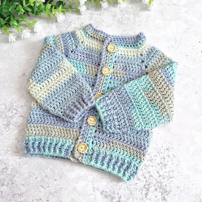 A handmade crocheted cardigan sweater for 0-3 months baby, featuring soft pastel stripes of light blue, mint green, and beige, with wooden buttons. It is laid flat on a light gray textured surface with decorative green leaves and white flowers in the top left corner.
