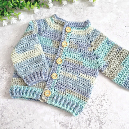 A handmade crocheted baby cardigan with horizontal stripes in light blue, mint green, and beige, featuring wooden buttons down the front. The cardigan is laid flat on a light textured surface with small green leaves and white flowers in the top left corner.