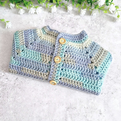 A handmade crocheted baby cardigan with horizontal stripes in light blue, mint green, and beige, featuring wooden buttons down the front, laid flat on a light gray textured surface with white flowers at the top.