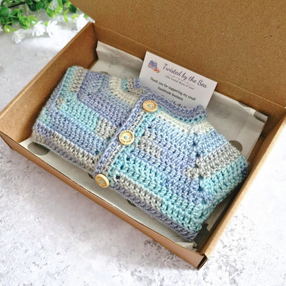 A handmade crocheted baby cardigan in pastel stripes of blue, purple, and beige, packaged inside a brown cardboard box with a thank you card.