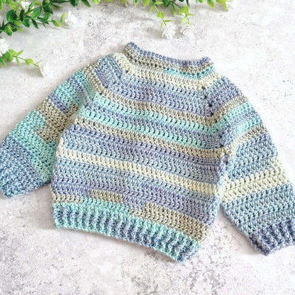 View of the back of a handmade crocheted sweater with horizontal stripes in light blue, mint green, and cream, laid flat on a textured light gray surface with green leaves in the top corners.