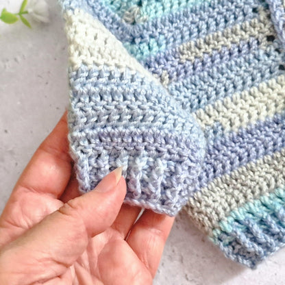A close-up of a hand holding the corner of a handmade crocheted Cardigan, showing its soft texture and striped pattern in pastel blue, lavender, and cream.