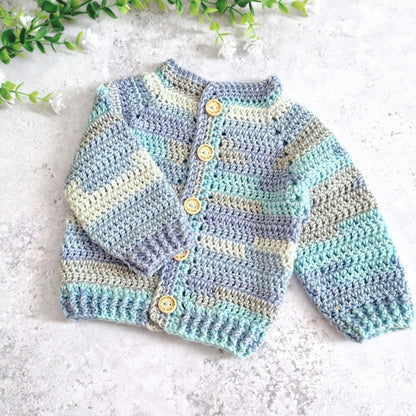 A handmade crochet cardigan sweater for babies, featuring horizontal stripes in light blue, mint green, and cream, with four wooden buttons down the front. It is laid flat on a textured light gray surface with green leaves in the top corners.