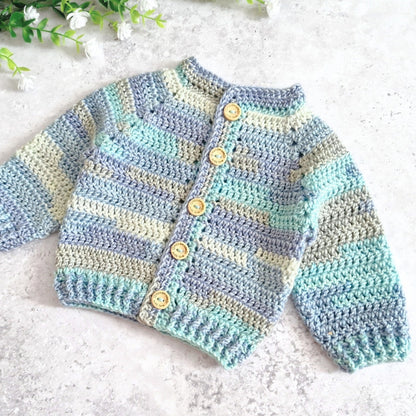 A handmade crocheted cardigan sweater with horizontal stripes in light blue, mint green, and cream, featuring five wooden buttons down the front, laid flat on a light gray textured surface with small white flowers in the corner.