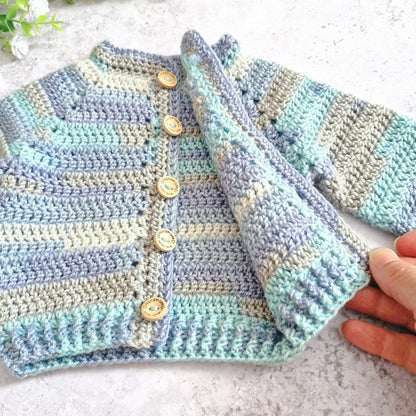A handmade crocheted cardigan with horizontal stripes in light blue, lavender, and cream, featuring wooden buttons and a ribbed collar, cuffs, and hem. A hand is holding up one of the sleeves to show the texture and drape of the yarn.