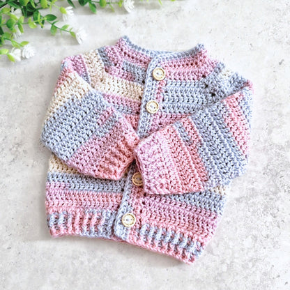 A handmade crocheted baby cardigan with pastel stripes of pink, light blue, and cream, featuring wooden buttons, laid flat on a light gray textured surface with small white flowers in the corner.