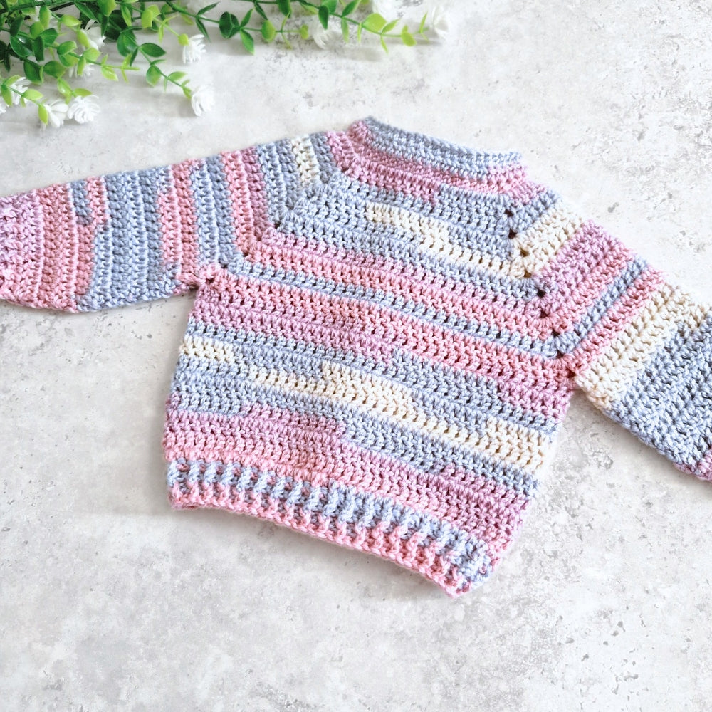Back view handmade crocheted baby sweater with horizontal stripes in pink, light blue, and cream, laid flat on a light gray textured surface with green leaves in the corner.