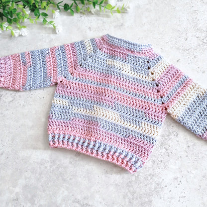 Back view handmade crocheted baby sweater with horizontal stripes in pink, light blue, and cream, laid flat on a light gray textured surface with green leaves in the corner.