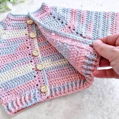 A close-up of a handmade crocheted cardigan with pink, light blue, and cream stripes, featuring wooden buttons, with a hand lifting one corner to show the texture.