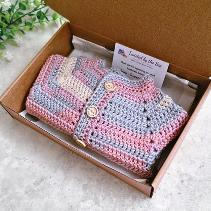 A handmade crocheted cardigan in pastel pink, light blue, and cream stripes, presented inside a brown cardboard gift box.