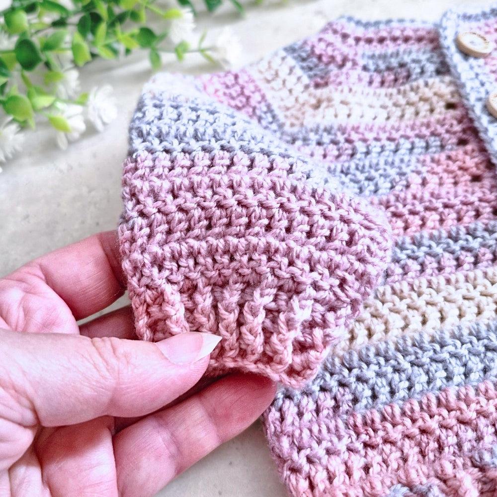 A close-up of a hand holding a pink textured crochet sleeve of a striped baby cardigan with pink, beige, and gray stripes.