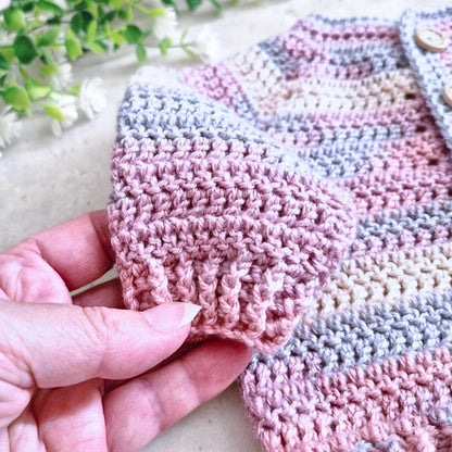 A close-up of a hand holding a pink textured crochet sleeve of a striped baby cardigan with pink, beige, and gray stripes.