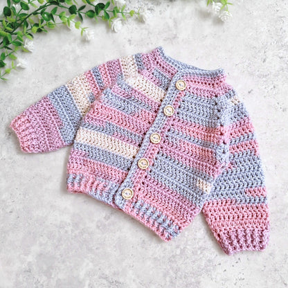 A handmade crocheted baby cardigan with horizontal stripes in pink, light blue, and cream, featuring wooden buttons down the front, laid flat on a light gray textured surface with green leaves in the corner. Yarn: King Cole Pebble Beach.