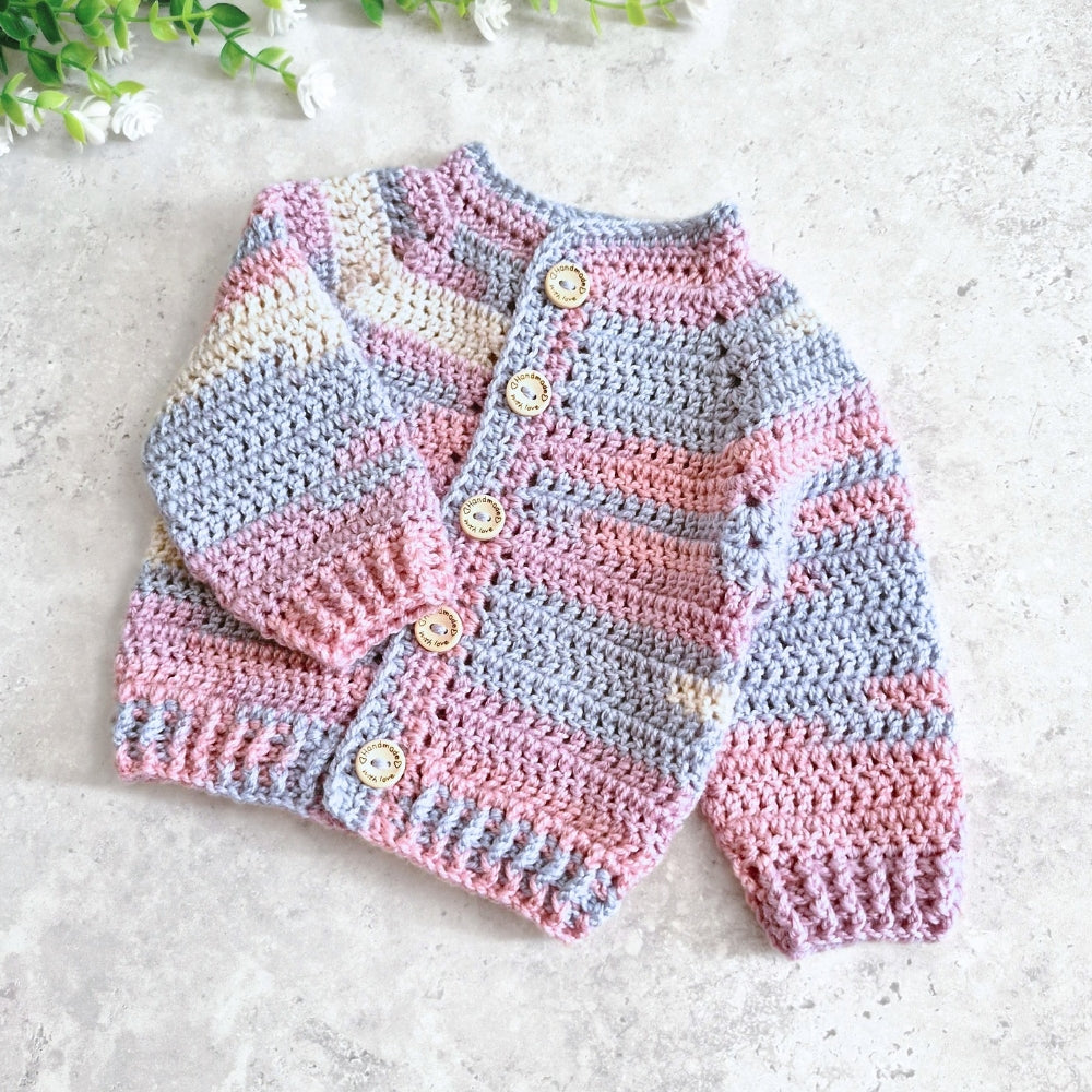 A handmade crocheted cardigan sweater for babies, featuring horizontal stripes in pink, light blue, and cream, with wooden buttons down the front, laid flat on a light gray textured surface.