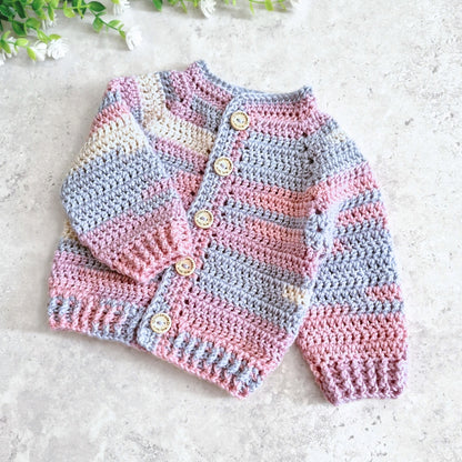 A handmade crocheted cardigan sweater for babies, featuring horizontal stripes in pink, light blue, and cream, with wooden buttons down the front, laid flat on a light gray textured surface.