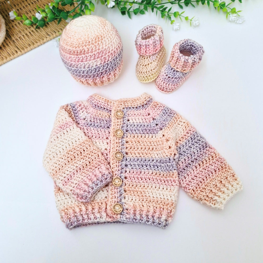 A handmade crocheted baby outfit set laid out on a white surface, including a long-sleeve cardigan with wooden buttons, a matching beanie, and a pair of booties. The items are made in soft pastel stripes of cream, pink, peach, and lavender-gray, with small green leaves and a woven mat in the background.