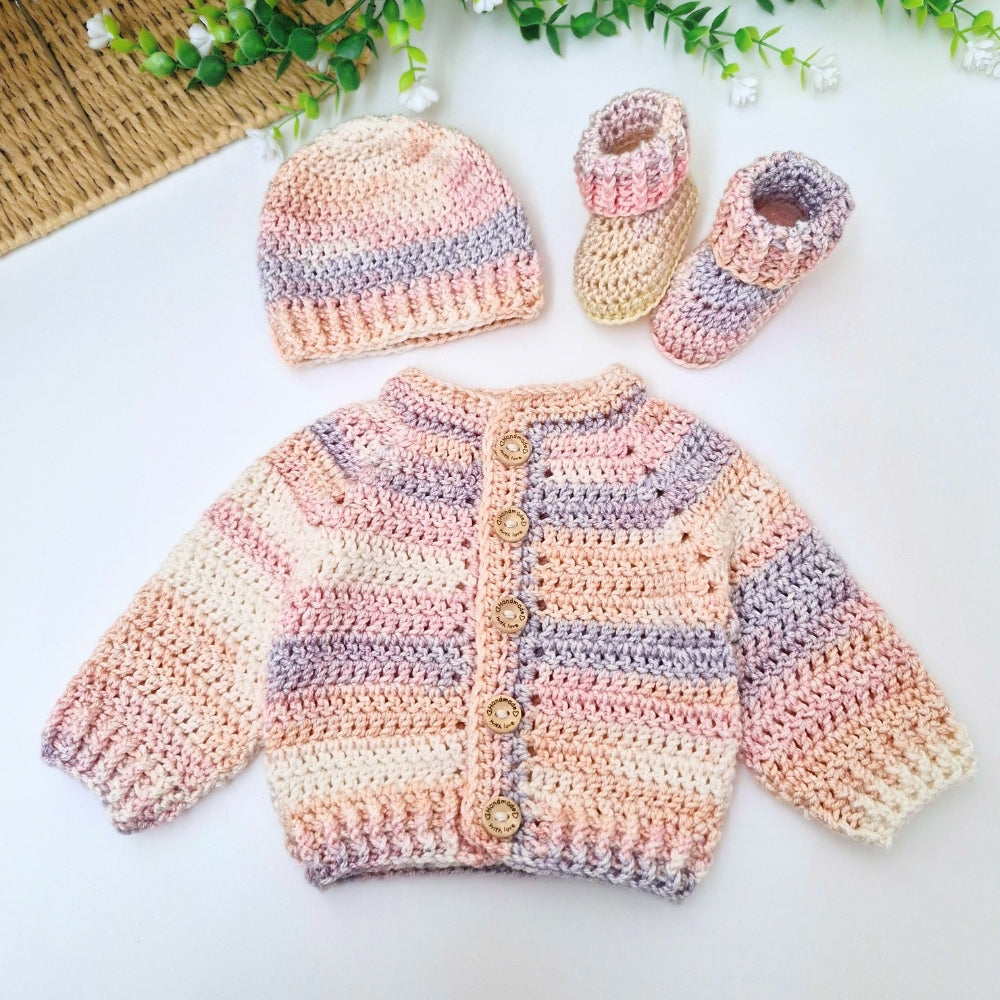 A handmade crocheted baby outfit set including a cardigan, a beanie, and booties, displayed on a white surface with decorative green leaves and a woven mat in the background.