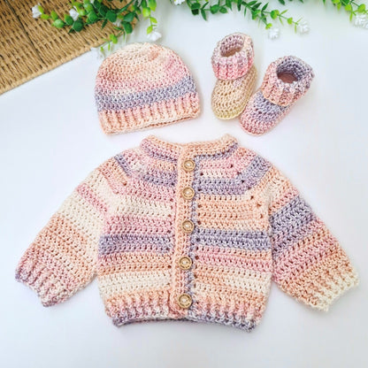 A handmade crocheted baby outfit set including a cardigan, a beanie, and booties, displayed on a white surface with decorative green leaves and a woven mat in the background.