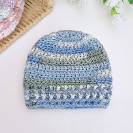 A handmade crocheted beanie hat featuring horizontal stripes of light blue, dusty blue, sage green, and white, displayed on a clean white surface with decorative props.