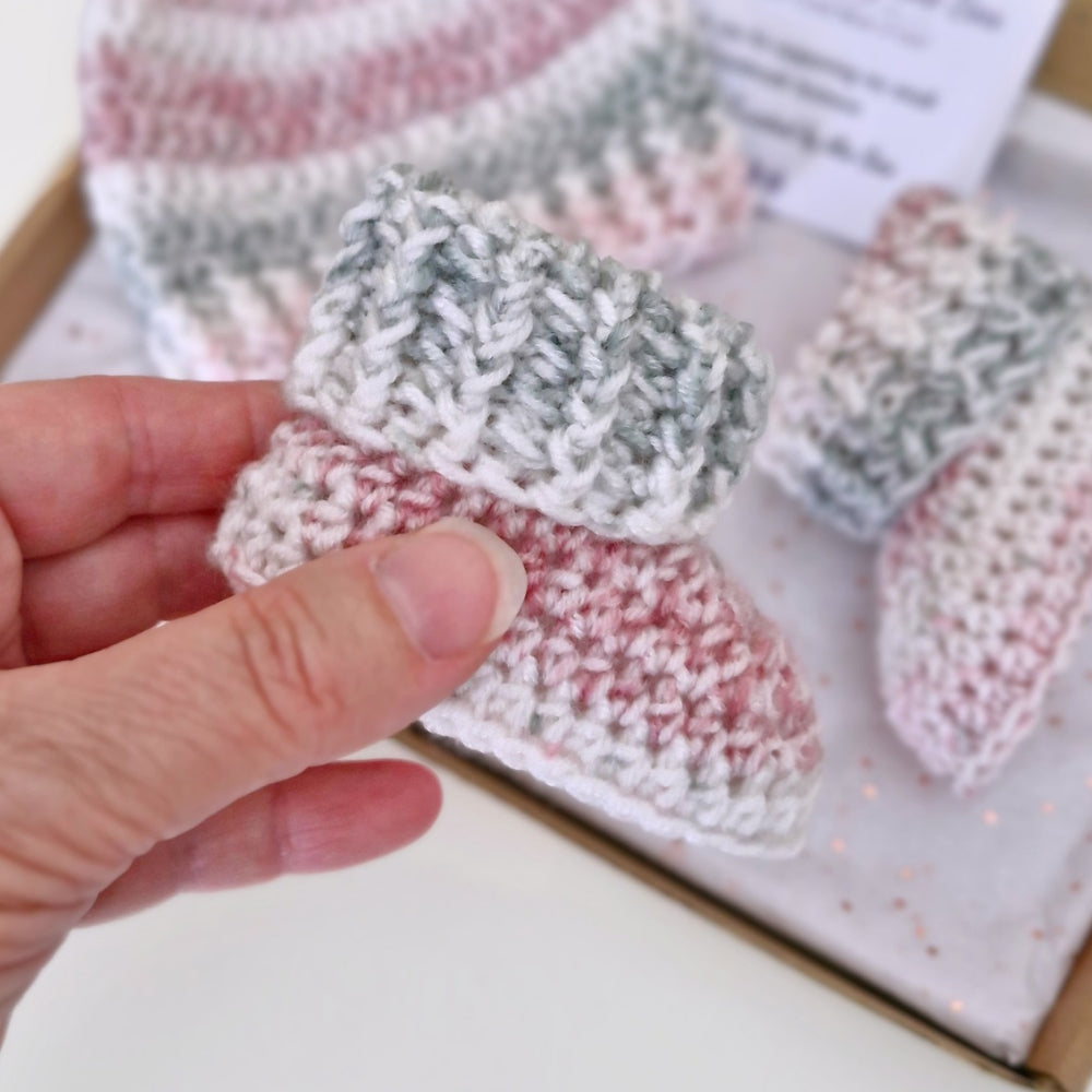 A close-up of a hand holding a crocheted baby bootie in pink, white, and gray marled yarn, with matching booties in the background inside a box.