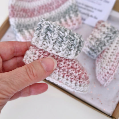 A close-up of a hand holding a crocheted baby bootie in pink, white, and gray marled yarn, with matching booties in the background inside a box.