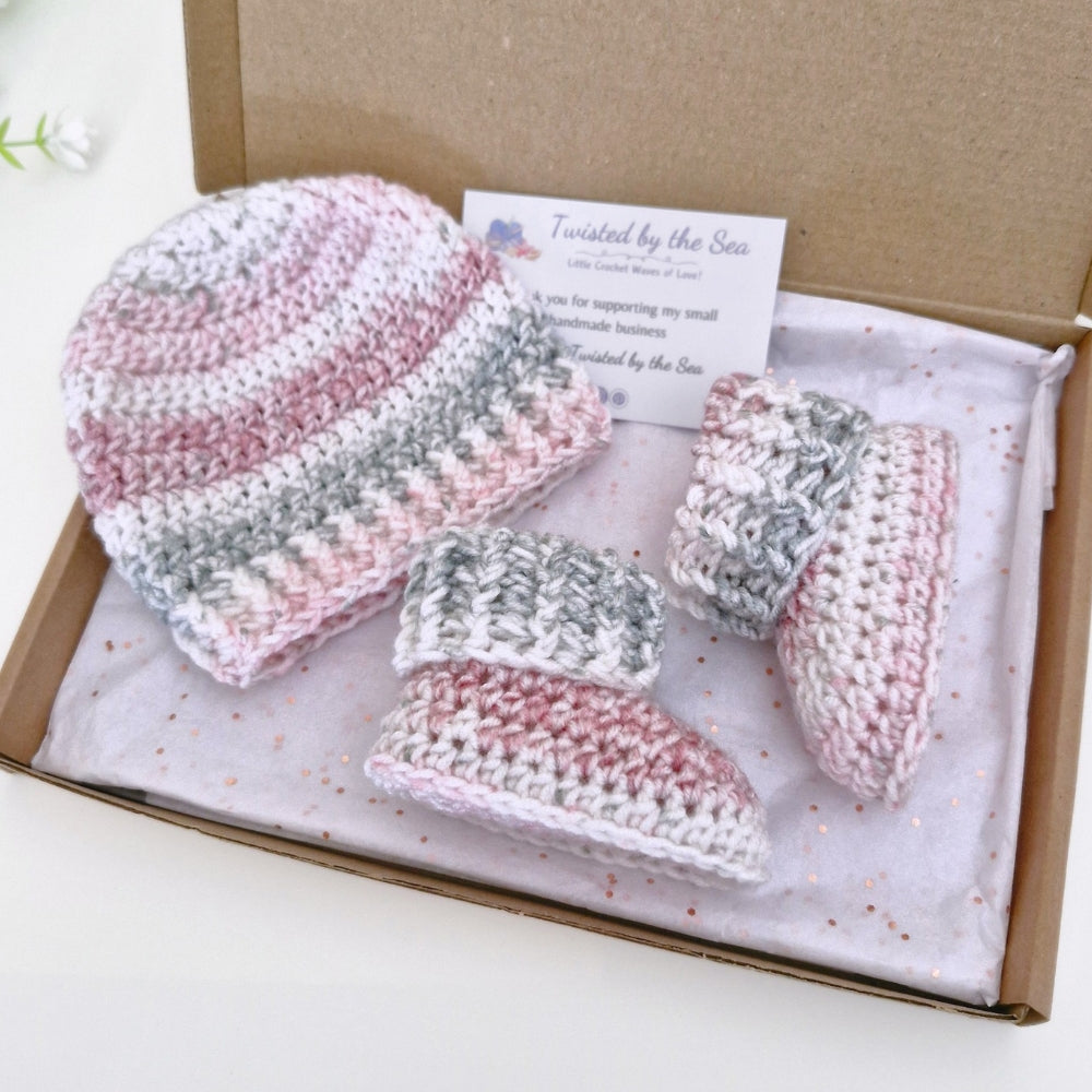 A handmade crochet baby gift set by Twisted by the Sea, including a beanie, booties, displayed inside a brown cardboard gift box with a white tissue paper and brand card.