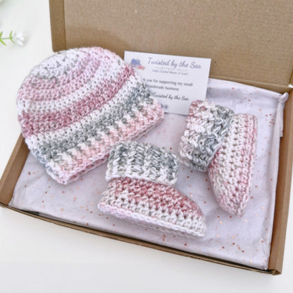 A handmade crochet baby gift set by Twisted by the Sea, including a beanie, booties, displayed inside a brown cardboard gift box with a white tissue paper and brand card.