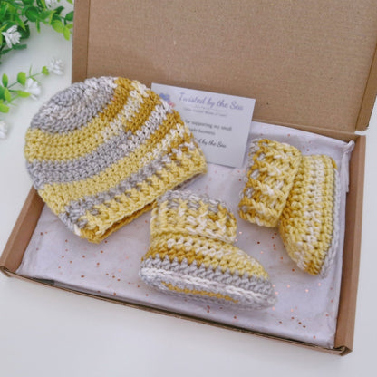 A handmade crochet baby bootie set in yellow, gray, and white stripes, presented in a brown cardboard gift box with a white tissue paper lining and a small card.