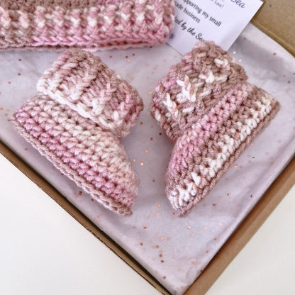A set of hand-crocheted baby booties and a matching hat in variegated pink, beige, and white yarn, displayed inside a gift box with tissue paper.