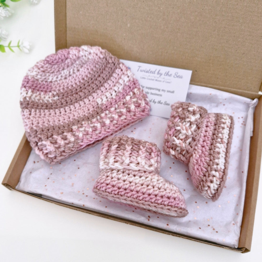 A handmade crochet baby hat and bootie set in variegated pink and beige yarn, presented inside a brown gift box with a tissue paper insert and a small card.