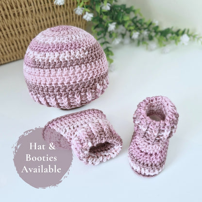 A crocheted baby hat and matching booties in soft pink and mauve stripes, arranged on a white surface with a wicker basket and greenery in the background. A circular badge reads 'Hat & Booties Available'.