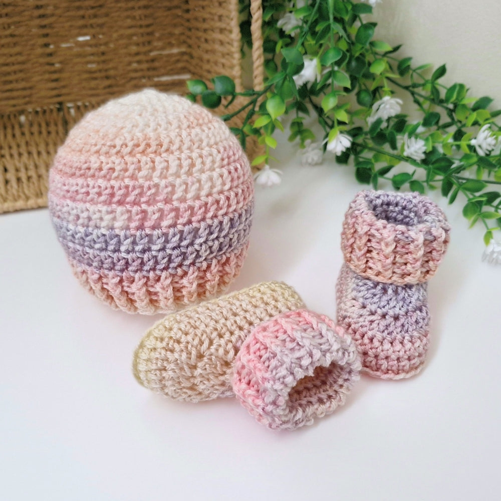A handmade crochet baby bootie set with a matching hat, displayed on a white surface with a wicker basket and green foliage in the background. The crochet items feature a soft pastel gradient of pink, beige, and lavender.